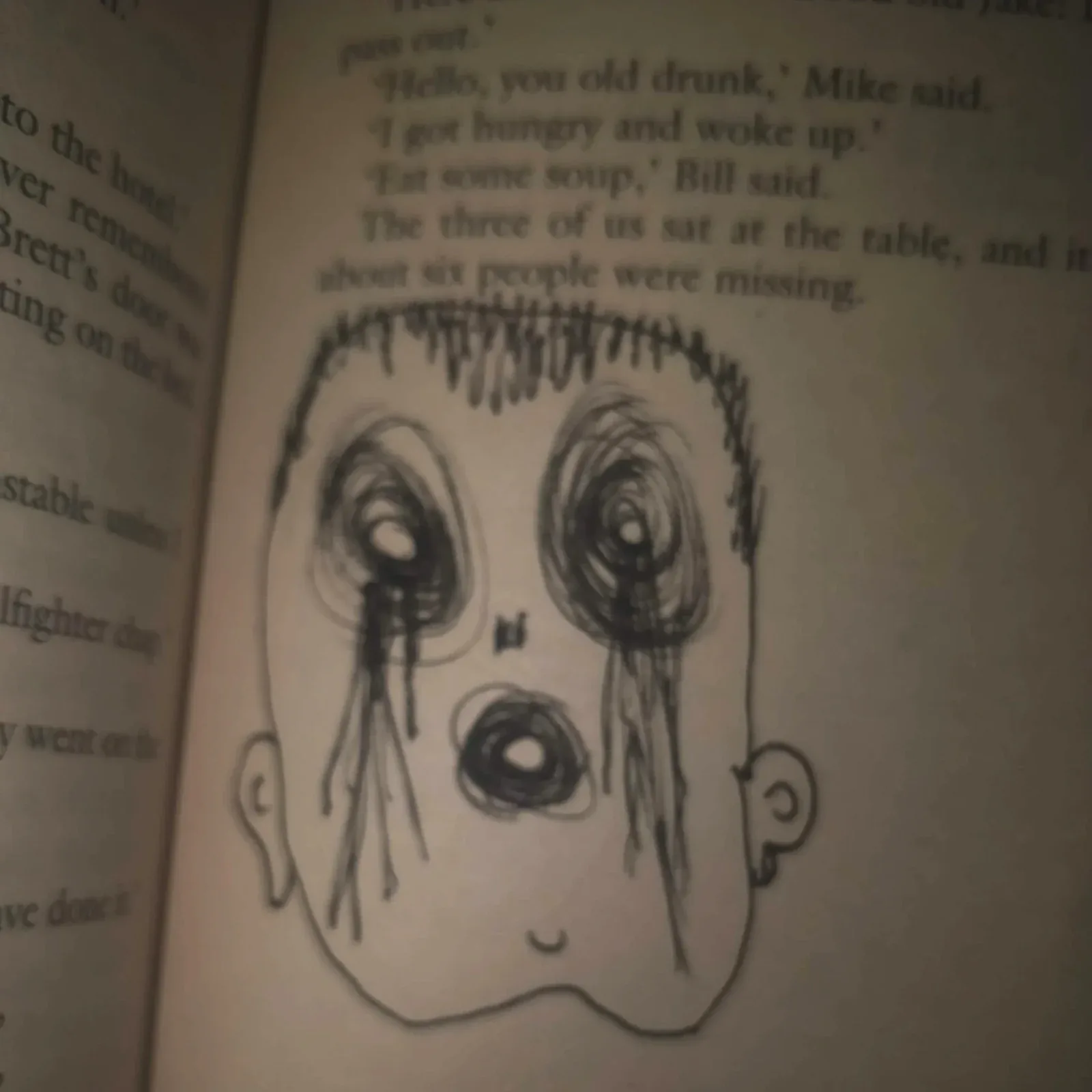 A face with hollow eyes drawn in pen over the printed text of a paperback