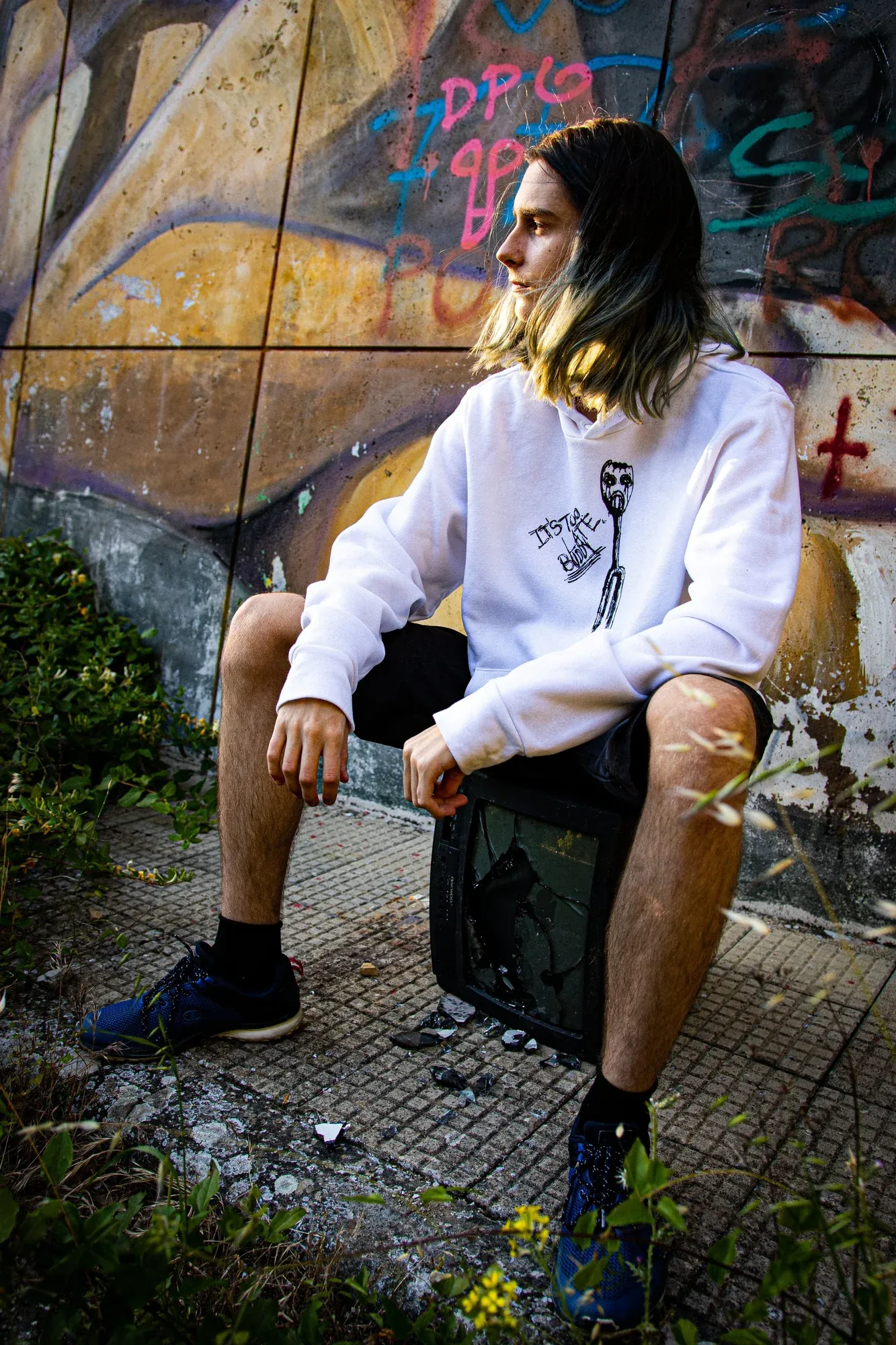 A man in a white BAKAGI hoodie sitting against a wall of graffiti