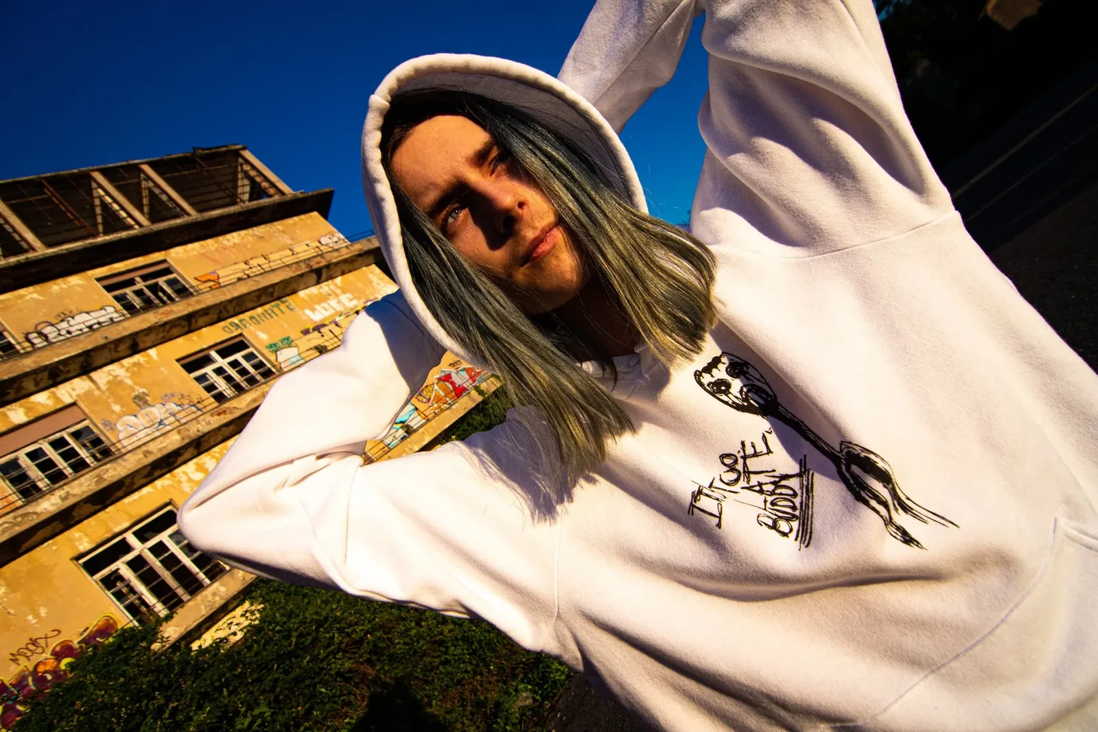 A man in a white hoodie reaching towards the camera, a ruined building behind him