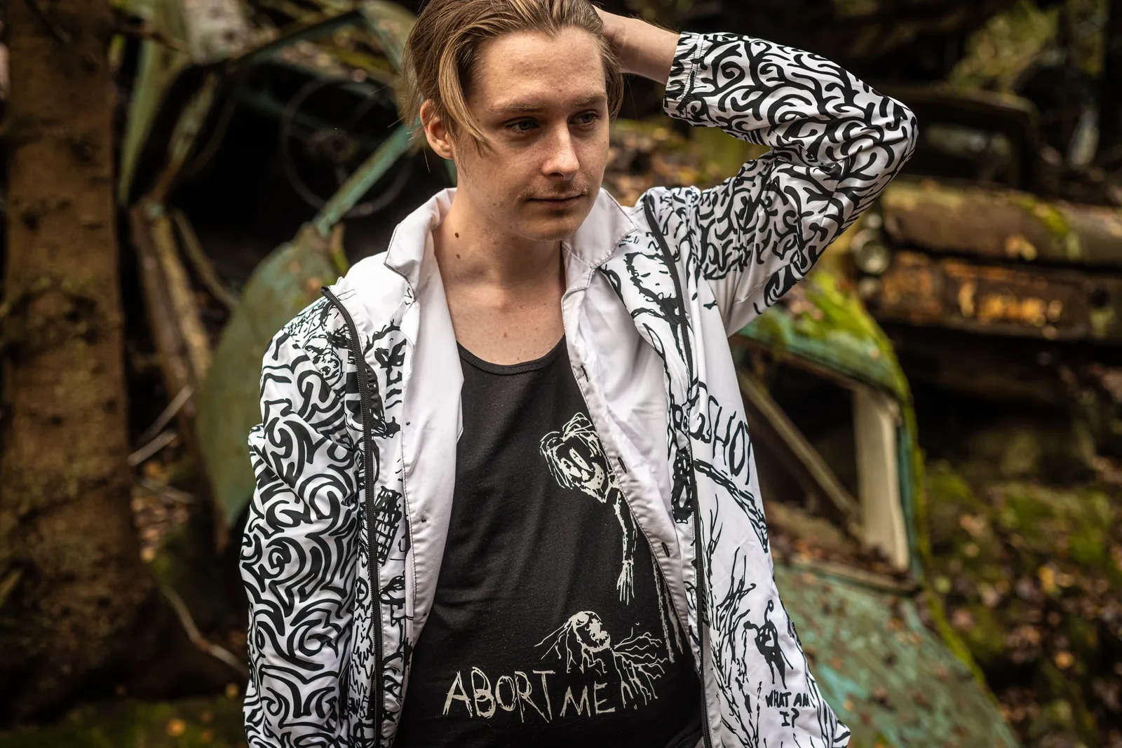 A young man in the Infinity windbreaker over the Abort Me tank top, leaning on a rusted car in the forest