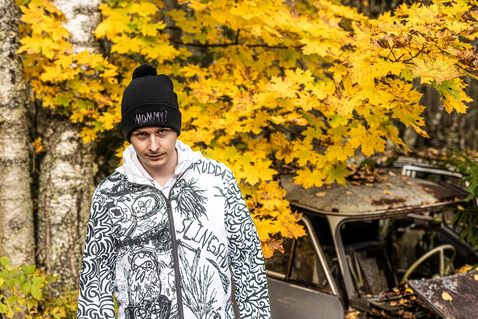 A young man in the Mommy? beanie and a doodled jacket under yellow autumn leaves