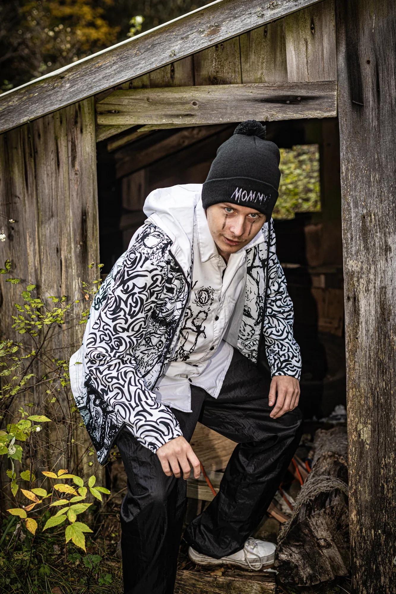A young man in the Mommy? beanie and a doodled jacket stepping out of a wooden shed