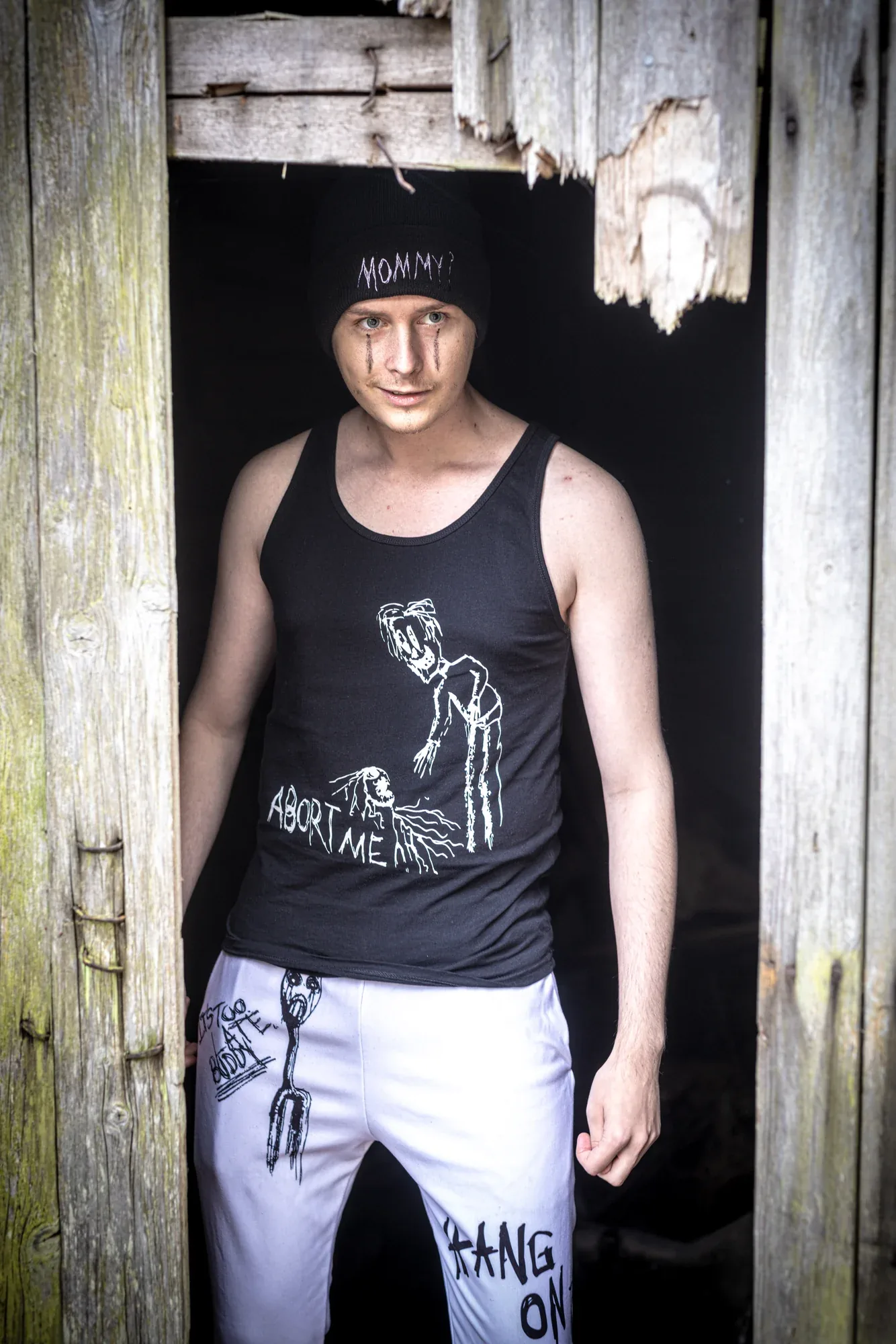 A young man in the Abort Me tank top and Hang On sweatpants in the doorway of a wooden shed