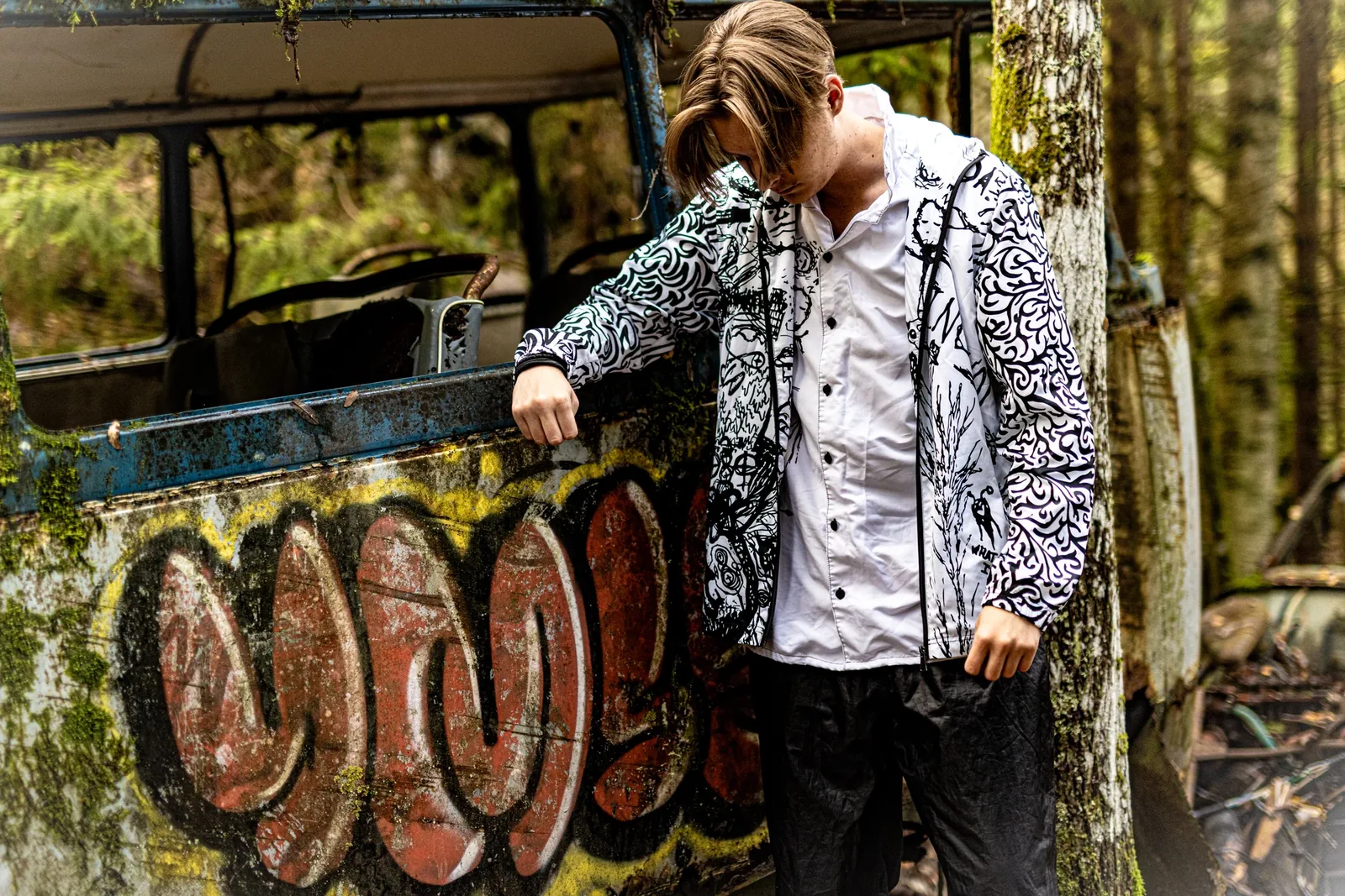 A young man in a doodled jacket leaning on a graffiti-covered bus in the forest